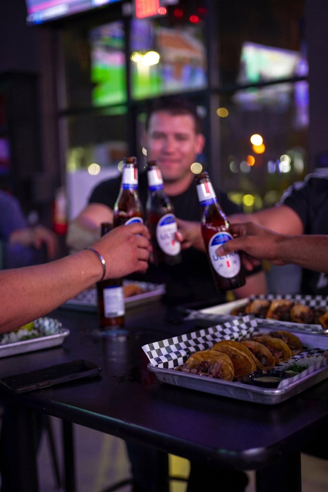 Friends toasting with beer bottles over a tray of tacos at SKYBOX