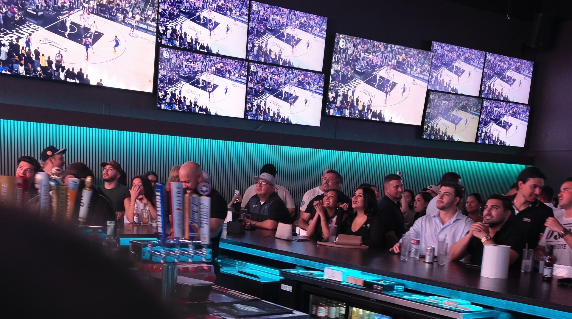 A packed SKYBOX bar under a row of TV screens, the bar glowing teal