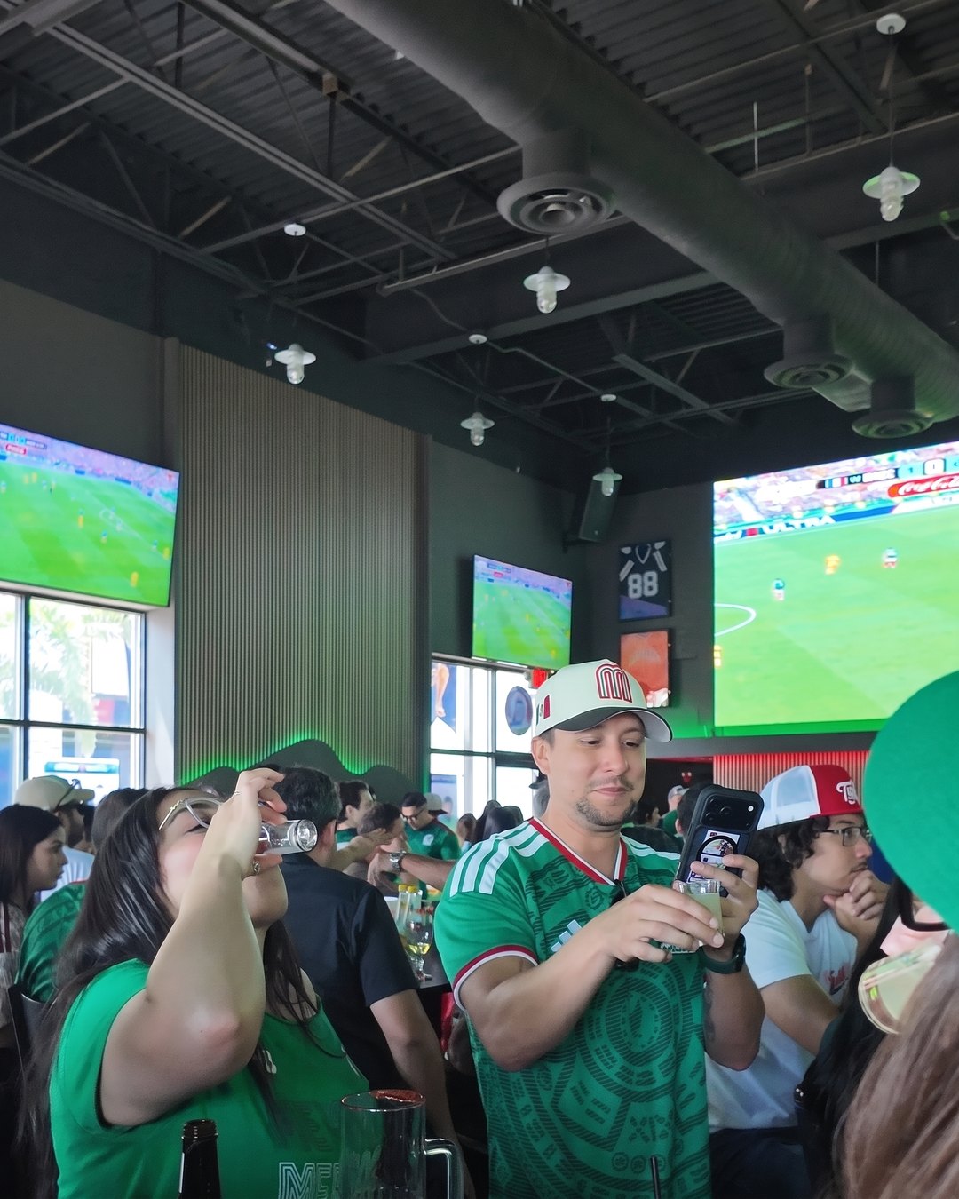 Fans in Mexico jerseys raising shots under big screens showing a World Cup match