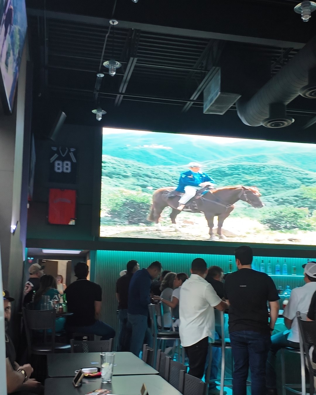 The Wall at SKYBOX: a giant LED screen over the teal-lit bar, guests standing at the bar below