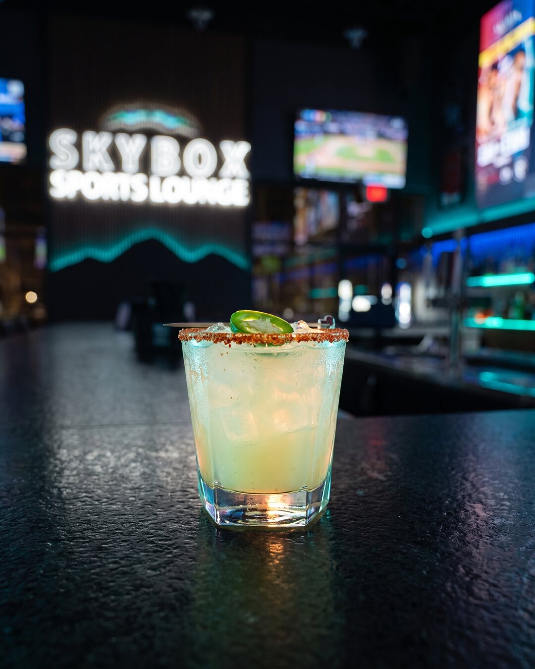 A chile-rimmed Skybox margarita with a jalapeño slice on the bar, the SKYBOX sign glowing behind